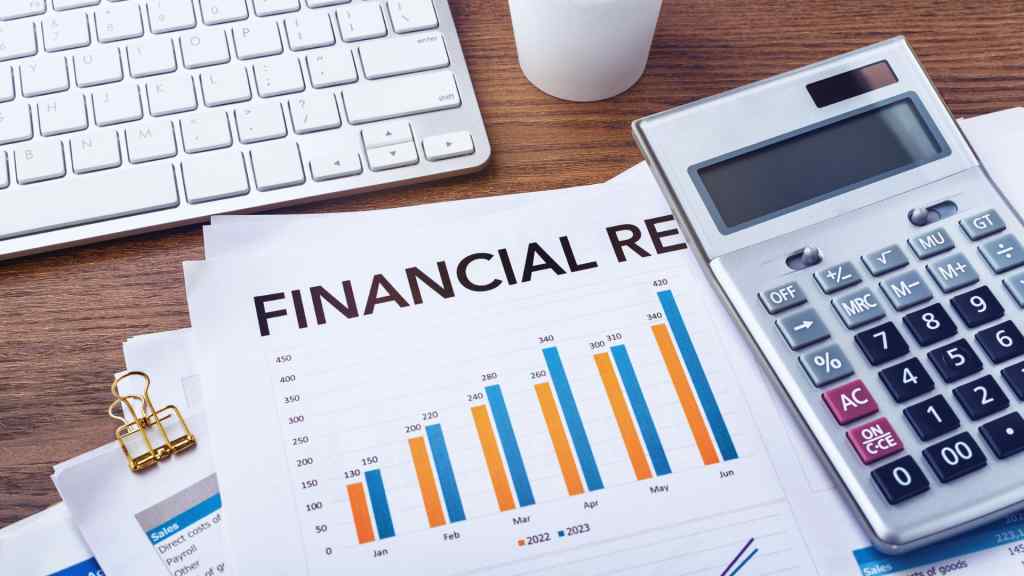 Alt text: A financial report with bar graphs lies on a wooden desk. Nearby are a calculator, a keyboard, gold paperclips, and a white cup, conveying a business setting.